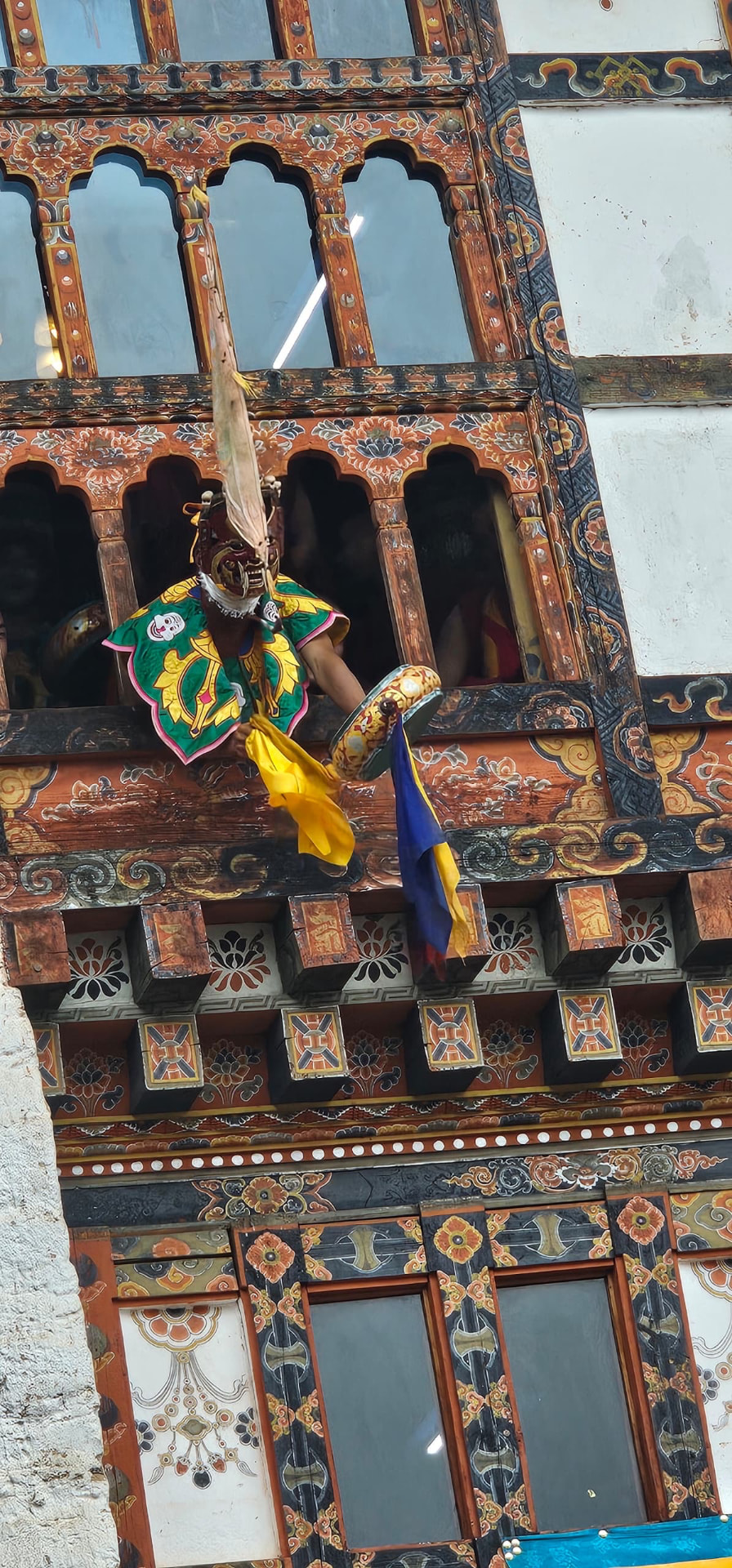 A masked performer in colourful costume beside intricately painted Bhutanese wooden windows
