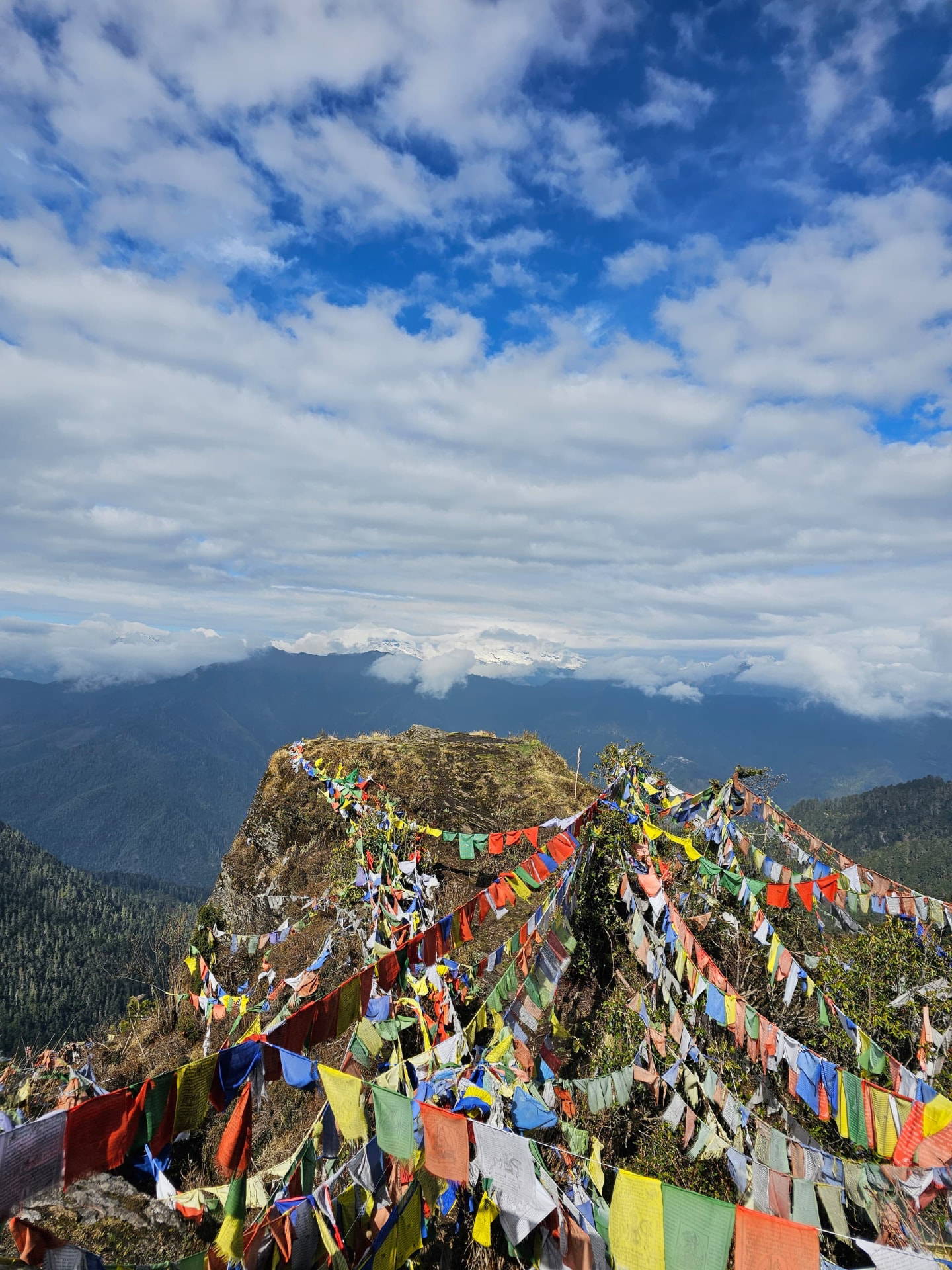 Prayer flags crowning a rocky mountain summit, with snow-capped peaks emerging from the clouds beyond