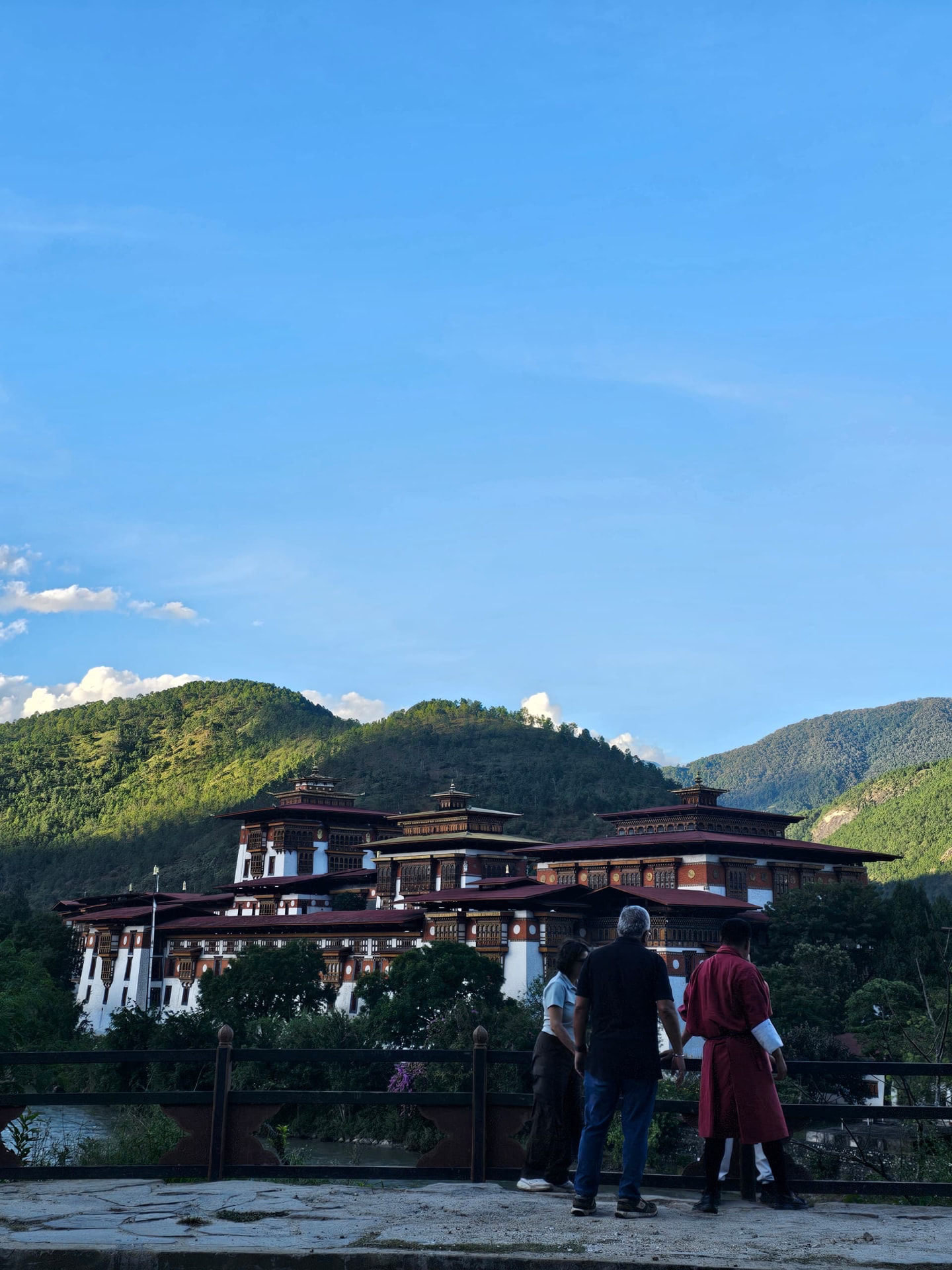 A large Bhutanese dzong beside a river, backed by forested hills under a broad blue sky