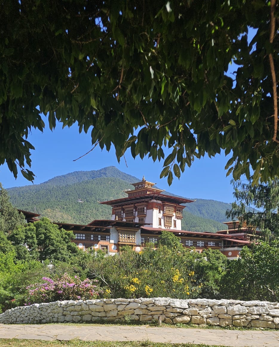 A Bhutanese dzong framed by leafy branches, with a stone wall and forested mountain behind