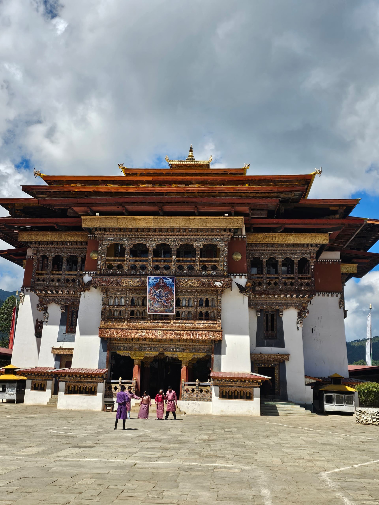 An ornate Bhutanese temple with gilded woodwork and visitors standing in its stone courtyard