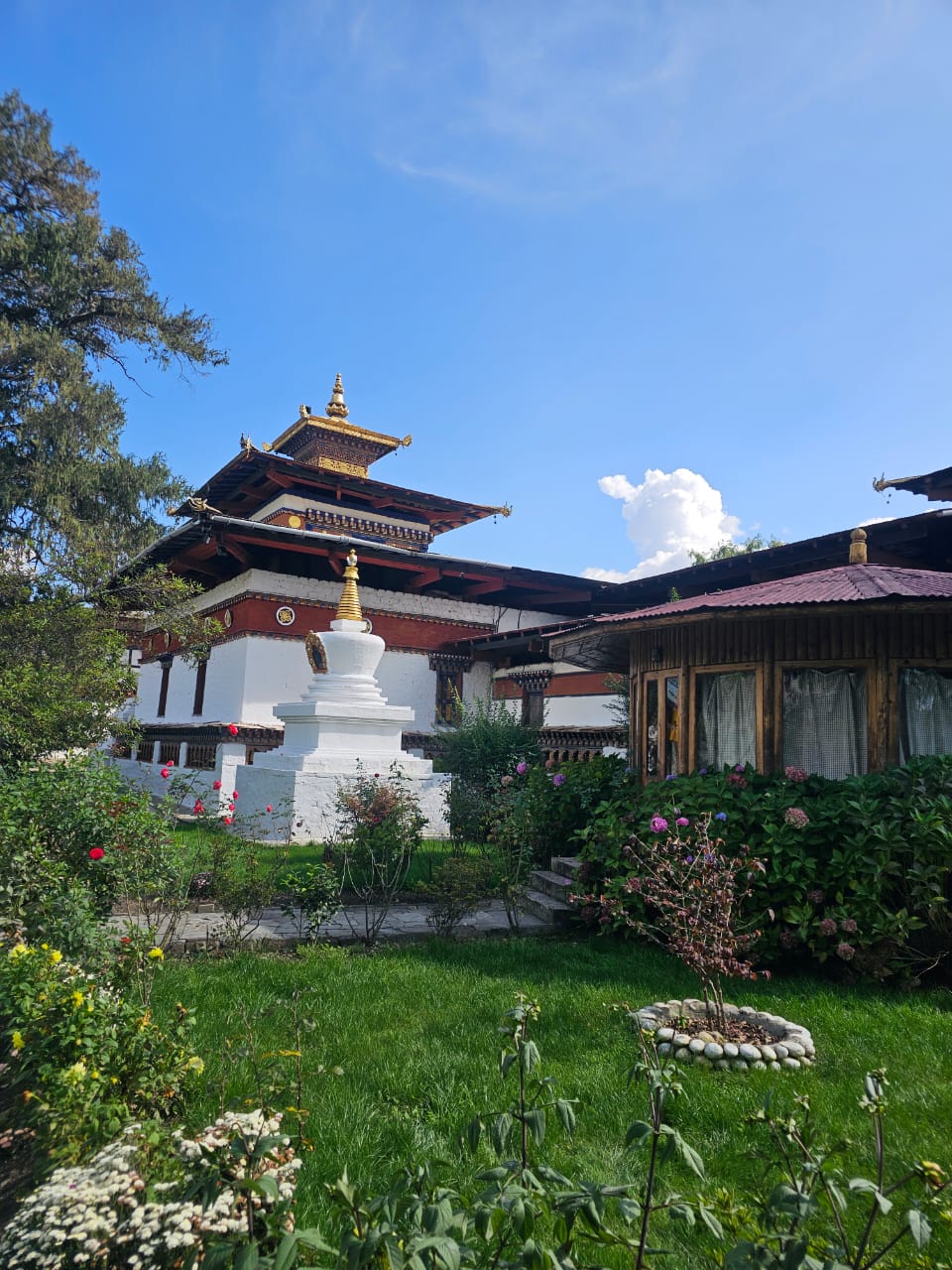 Kyichu Lhakhang in the Paro Valley — a whitewashed temple with tiered wooden roofs and a golden finial beside a white chorten, standing in a flower-filled garden lawn