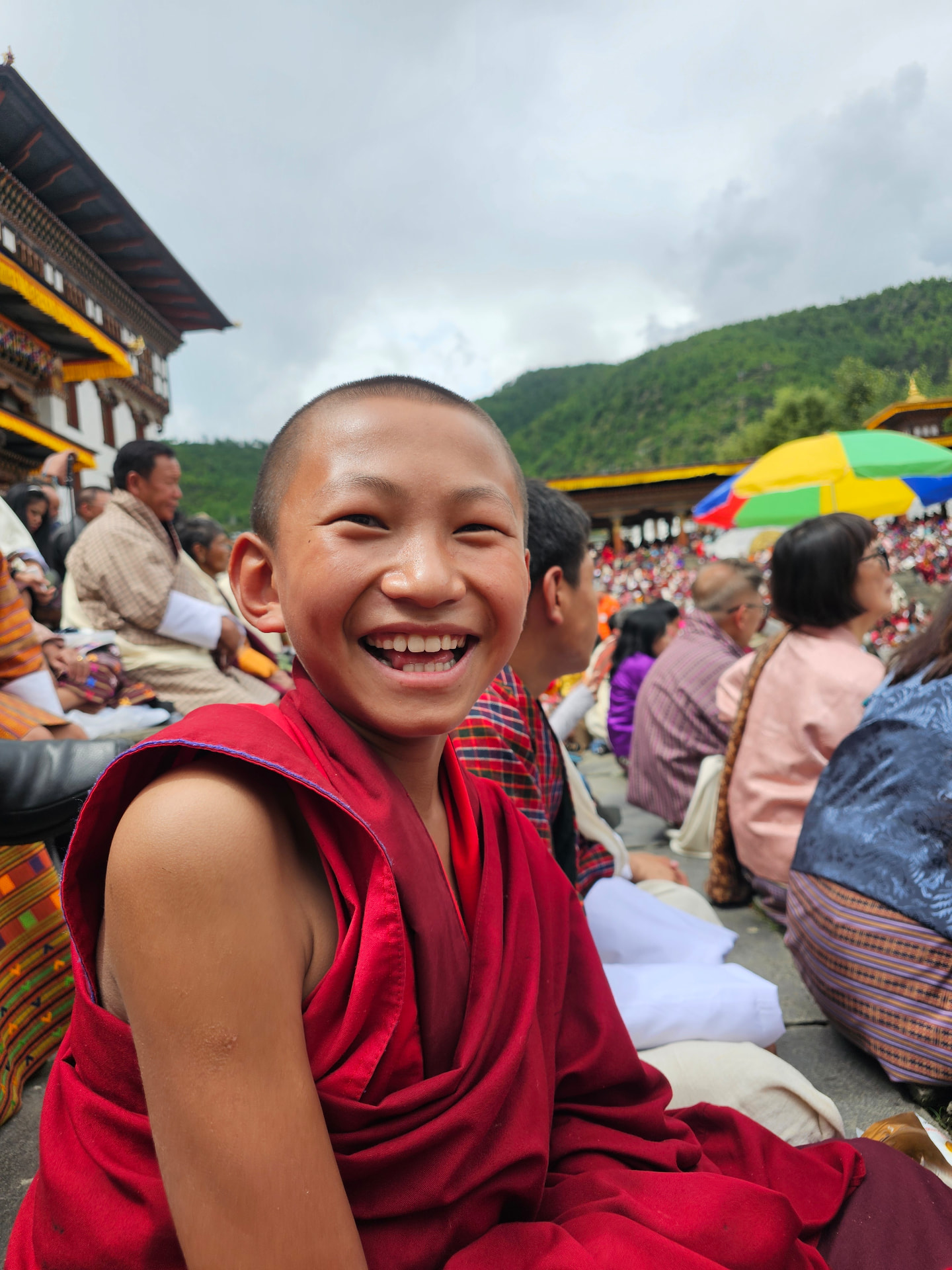 A smiling young monk in a red robe seated among festivalgoers