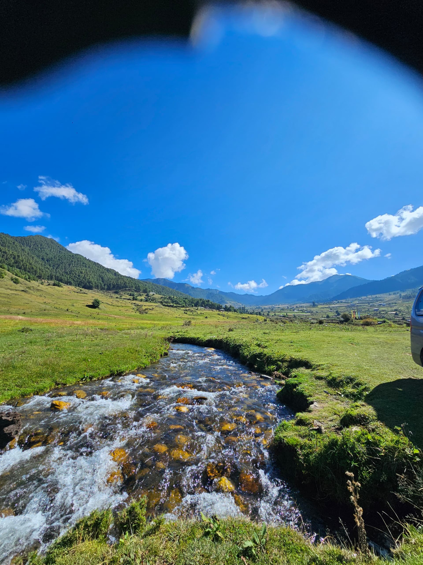 A grassy Himalayan mountain valley with a stream running through it under a bright sky