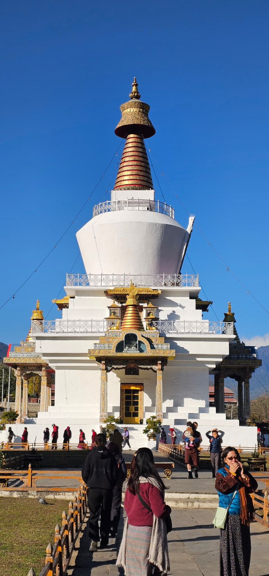 A white and gold Buddhist chorten beneath a clear blue sky, with visitors circling in the foreground